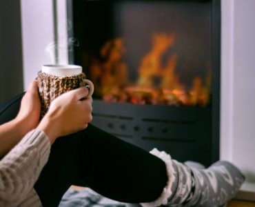 Young girl sitting in front of the fireplace and holding cup of tea in hand on legs and warming. Woman in winter clothes and wool socks. Winter and cold weather concept. Close up, selective focus