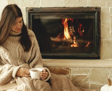 Woman with cup of warm tea relaxing at fireplace, autumn hygge. Heating house with wood burning stove. Young stylish female in cozy sweater sitting at fireplace in rustic farmhouse