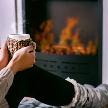 Young girl sitting in front of the fireplace and holding cup of tea in hand on legs and warming. Woman in winter clothes and wool socks. Winter and cold weather concept. Close up, selective focus