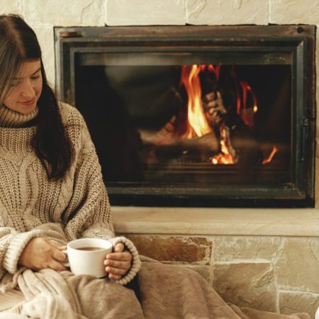 Woman with cup of warm tea relaxing at fireplace, autumn hygge. Heating house with wood burning stove. Young stylish female in cozy sweater sitting at fireplace in rustic farmhouse